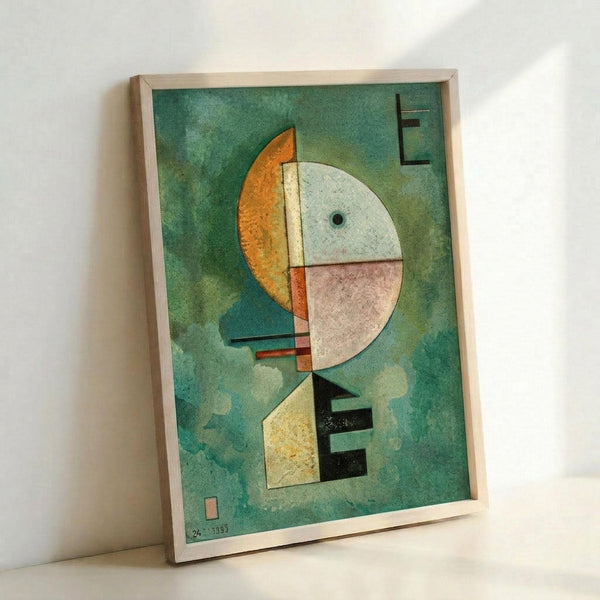 Abstract modernist artwork with geometric shapes on green watercolor background. Features a large circular form divided into orange, white, and pink sections with a small black dot, positioned above angular geometric forms in yellow, black, and green. Rectangular elements marked with "E" appear in corners. Displayed in light wood frame leaning against white wall.