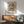 May include: Van Gogh roses poster print in light wood frame hanging on white wall above walnut credenza. Artwork shows white and pink roses in vase with impressionist brushstrokes against rose-colored background. Mid-century modern room setting with sage green armchair, small table lamp, and natural sunlight casting geometric shadows across the scene.