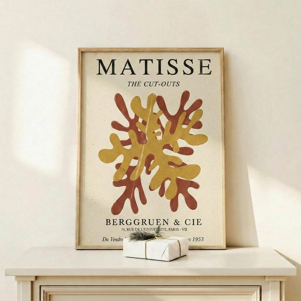 May include: Matisse cut-outs exhibition poster in light wood frame leaning against white wall on natural wood shelf. Features abstract coral and golden yellow organic shapes resembling sea anemones or coral formations on cream background. Text reads "MATISSE THE CUT-OUTS" and "BERGGRUEN & CIE" with Paris gallery details. Small wrapped gift with evergreen sprig sits beside frame.
