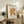 May include: Framed Matisse exhibition poster with abstract coral-like cut-out design in golden yellow and rust red colors, displayed on rustic wooden sideboard alongside fern plant, ceramic vases, and stacked books in warm-toned living room setting with cream upholstered furniture and soft natural lighting.