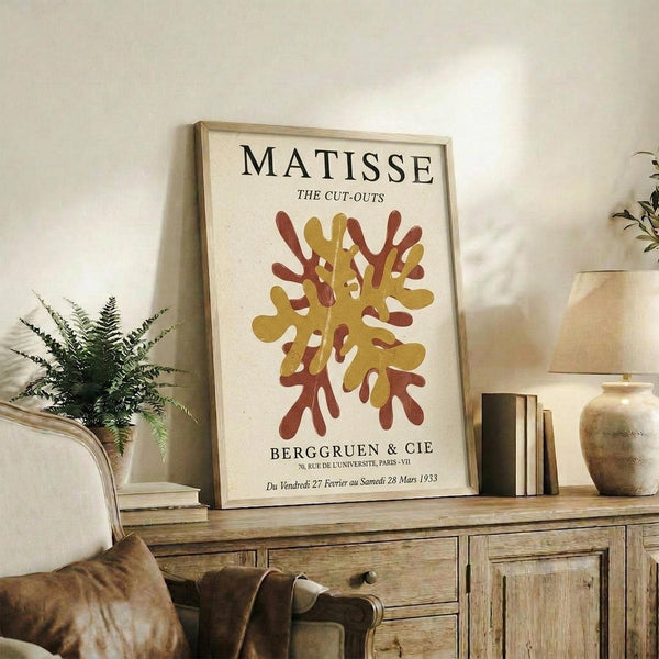 May include: Framed Matisse exhibition poster with abstract coral-like cut-out design in golden yellow and rust red colors, displayed on rustic wooden sideboard alongside fern plant, ceramic vases, and stacked books in warm-toned living room setting with cream upholstered furniture and soft natural lighting.