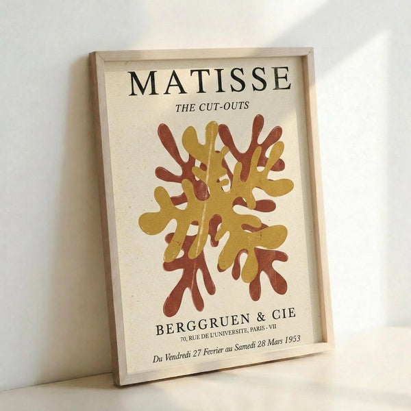May include: Framed art print featuring abstract coral-like shapes in golden yellow and rust red colors on cream background. Text reads "MATISSE THE CUT-OUTS" at top and "BERGGRUEN & CIE" with French exhibition details at bottom. Light wood frame displayed against white wall, casting soft shadow.