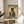 May include: Framed vintage-style poster featuring a black cat in traditional Japanese clothing holding a red coffee cup, displayed on a wooden round stool next to a beige upholstered chair. The artwork has aged parchment background with Japanese characters, green text banners reading "MORE ESPRESSO" and "LESS DEPRESSO." Natural sunlight streams across the neutral-toned room setting with a plant visible in the corner.