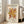 May include: Framed vintage-style exhibition poster featuring abstract coral-like shapes in golden yellow and rust red colors on cream background. The artwork shows organic, flowing forms reminiscent of sea coral or algae. Black text appears at top and bottom with gallery information and French exhibition dates from 1953. The wooden-framed poster leans against a white wall next to a beige upholstered chair in a bright, minimalist room with large windows.