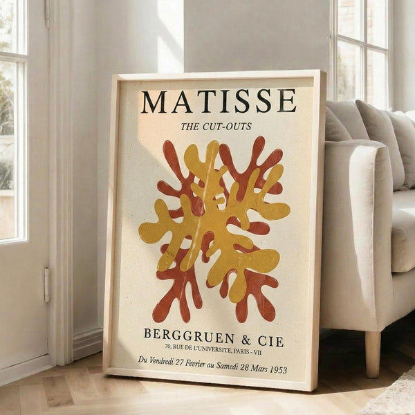 May include: Framed vintage-style exhibition poster featuring abstract coral-like shapes in golden yellow and rust red colors on cream background. The artwork shows organic, flowing forms reminiscent of sea coral or algae. Black text appears at top and bottom with gallery information and French exhibition dates from 1953. The wooden-framed poster leans against a white wall next to a beige upholstered chair in a bright, minimalist room with large windows.