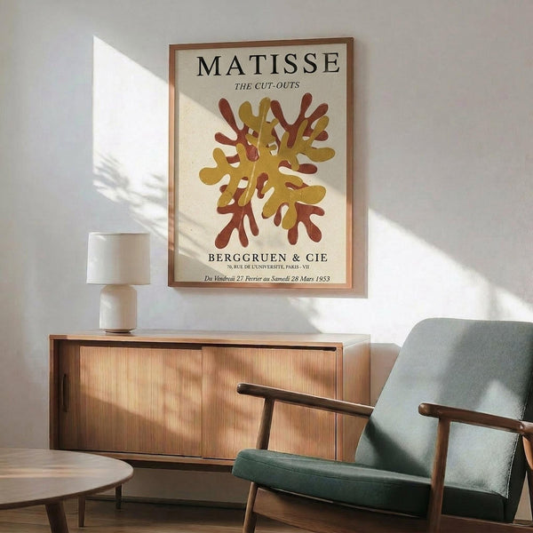 May include: Framed Matisse exhibition poster featuring abstract cut-out shapes in golden yellow and rustic red tones, displayed above a mid-century modern wooden credenza with white ceramic lamp. Sage green upholstered armchair with wooden frame sits nearby on light wooden floors. Warm sunlight creates diagonal shadows across white walls in this minimalist living room setting.