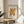 May include: Framed vintage exhibition poster featuring abstract coral-like cutout design in golden yellow and rust red colors, displayed on round wooden side table next to beige upholstered chair, with plant leaves visible and natural sunlight casting shadows across cream-colored wall