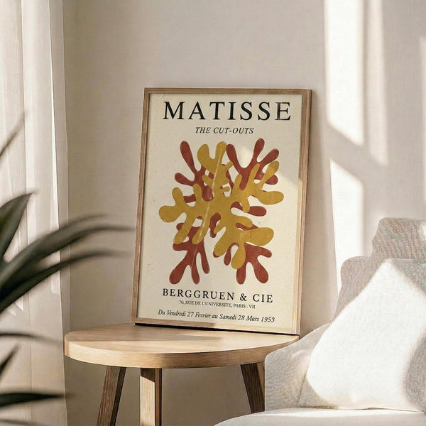 May include: Framed vintage exhibition poster featuring abstract coral-like cutout design in golden yellow and rust red colors, displayed on round wooden side table next to beige upholstered chair, with plant leaves visible and natural sunlight casting shadows across cream-colored wall