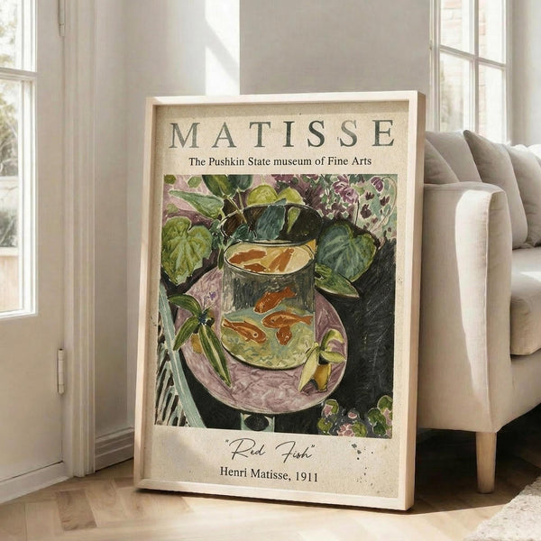 May include: Framed museum poster featuring Henri Matisse's colorful painting of red fish swimming in a glass fishbowl surrounded by lush green foliage and plants. The artwork displays vibrant greens, reds, and purples in Matisse's distinctive style. Light wooden frame leans against white wall next to beige upholstered chair in bright, airy room with large windows and natural lighting.