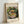 May include: Framed art print leaning against white wall showing Matisse's colorful painting of red goldfish in glass bowl surrounded by lush green plants. Print has cream matting with "MATISSE" and "The Pushkin State museum of Fine Arts" text at top, artwork title and "Henri Matisse, 1911" at bottom. Natural wood frame catches soft lighting creating gentle shadows.