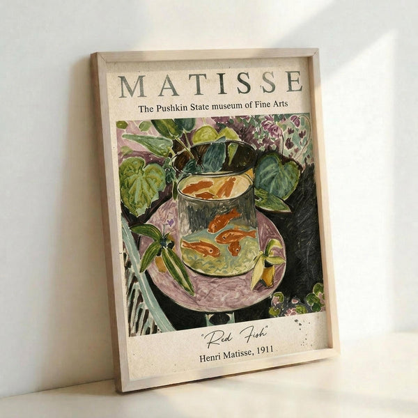 May include: Framed art print leaning against white wall showing Matisse's colorful painting of red goldfish in glass bowl surrounded by lush green plants. Print has cream matting with "MATISSE" and "The Pushkin State museum of Fine Arts" text at top, artwork title and "Henri Matisse, 1911" at bottom. Natural wood frame catches soft lighting creating gentle shadows.