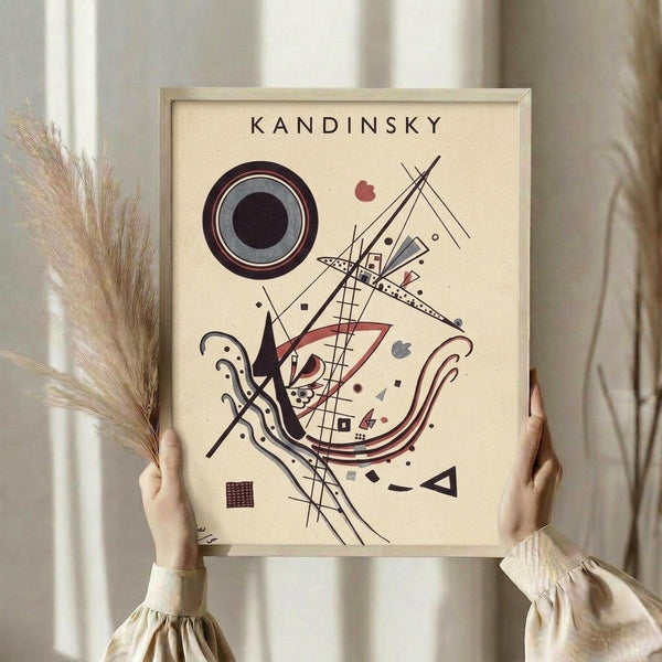 May include: Person holding framed abstract art print in cream-colored room setting with dried pampas grass. The artwork features geometric shapes, circles, lines and forms in black, red, and muted tones on cream background. Light wooden frame complements neutral aesthetic with soft natural lighting from window.