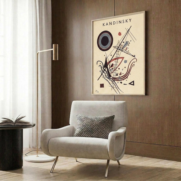May include: Framed Kandinsky abstract art print hanging on wood-paneled wall above cream upholstered armchair with geometric pillow. Print features black circles, red triangles, and intersecting lines on cream background. Modern brass floor lamp and dark side table with open book complete the warm, contemporary room setting.
