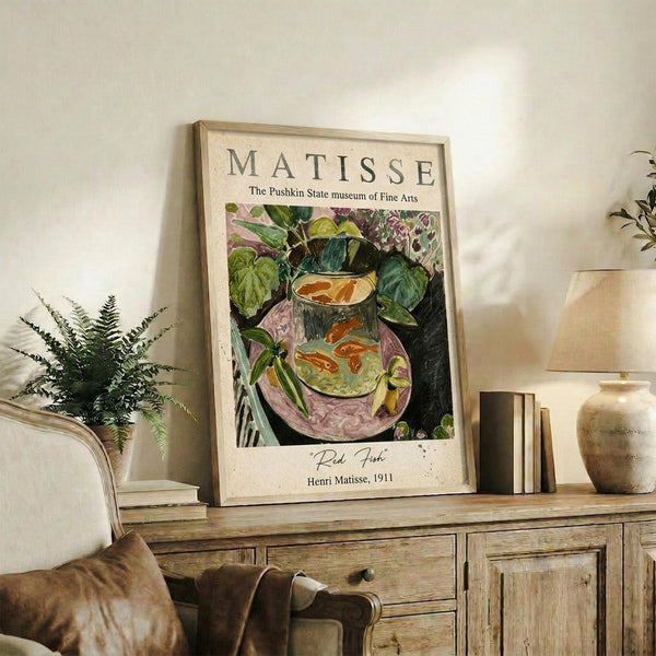 May include: Framed Matisse museum poster featuring a colorful still life of goldfish in a glass bowl surrounded by leafy plants, displayed on a rustic wooden sideboard next to a fern plant, ceramic vases, and books in a cozy living room setting with neutral tones and natural lighting