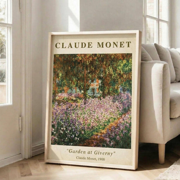 May include: Framed art print leaning against white wall next to beige sofa in bright room with windows. Print shows impressionist garden scene with purple and pink flowers in foreground, lush green trees overhead, and winding path. Text reads "CLAUDE MONET" at top and "Garden at Giverny" with "Claude Monet, 1900" at bottom. Light wood frame on hardwood floors with natural sunlight streaming in.