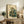 May include: Framed Japanese-style artwork of a green frog wearing traditional kimono and smoking a pipe, displayed on rustic wooden console with books, ceramic vase, and fern plant in neutral-toned living room setting with beige armchair visible