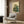 May include: Framed poster on wood-paneled wall showing a green frog in traditional Japanese kimono smoking a pipe, rendered in vintage ukiyo-e art style with Japanese characters. Modern beige armchair with geometric pillow sits below, alongside brass floor lamp and dark round side table with open book.