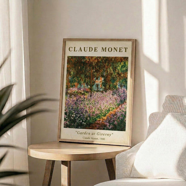 May include: Framed art print of Monet's Garden at Giverny displaying vibrant purple and pink flowers beneath green trees, propped on a light wood round side table next to a beige upholstered chair. Natural sunlight streams across the neutral-toned room, casting soft shadows from a plant's leaves visible in the corner.