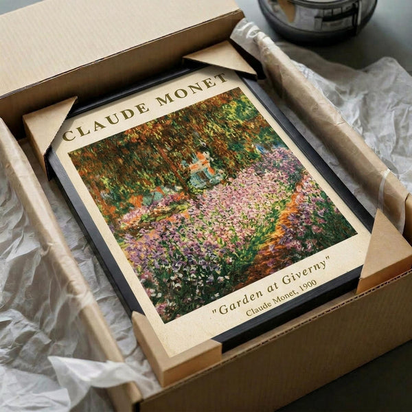May include: Framed art print of Claude Monet's impressionist garden painting featuring vibrant purple, pink and green flowering plants beneath drooping willow trees. The cream-colored print with black frame sits in an open cardboard shipping box with white tissue paper, next to wrapped cylindrical items on a neutral surface.