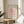 May include: Framed art print featuring a minimalist line drawing of a pink flamingo on a rose-colored background, displayed on a round wooden side table next to a beige upholstered chair. Natural sunlight streams across the neutral-toned room setting with a plant visible in the corner. The wooden frame complements the warm, modern interior styling.