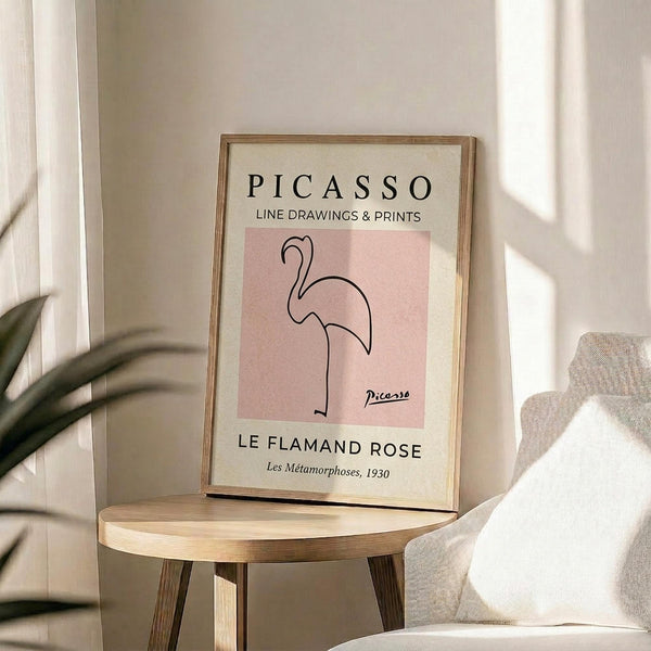 May include: Framed art print featuring a minimalist line drawing of a pink flamingo on a rose-colored background, displayed on a round wooden side table next to a beige upholstered chair. Natural sunlight streams across the neutral-toned room setting with a plant visible in the corner. The wooden frame complements the warm, modern interior styling.