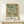 May include: Framed botanical poster featuring intricate green and sage foliage pattern with orange and cream flower clusters, displayed on light wood console table beside small wrapped gift with evergreen sprig against cream wall