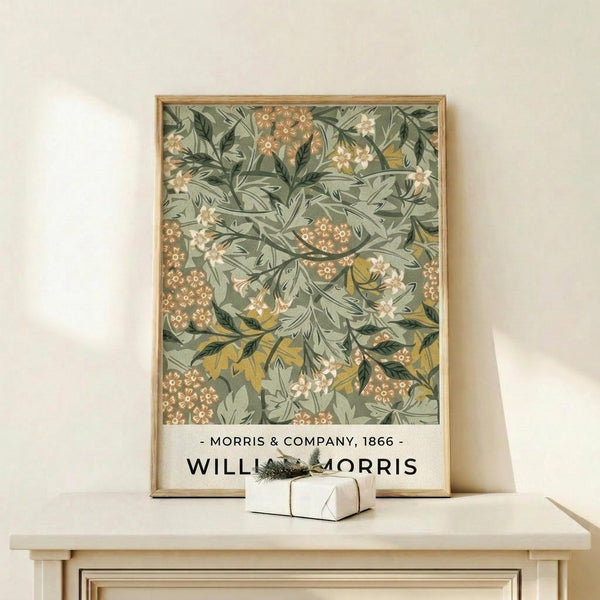 May include: Framed botanical poster featuring intricate green and sage foliage pattern with orange and cream flower clusters, displayed on light wood console table beside small wrapped gift with evergreen sprig against cream wall