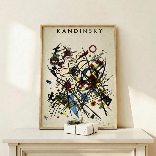 May include: Framed Kandinsky abstract art print displayed on light wood shelf against cream wall. The artwork features dynamic black lines, geometric shapes, and colorful elements in red, blue, yellow, and green on white background. Small wrapped gift box with evergreen sprig sits beside the thin wooden frame.