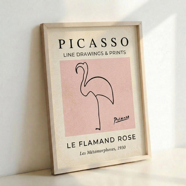 A framed art print leaning against a white wall, featuring a minimalist line drawing of a flamingo in black ink on a soft pink square background. The cream-colored poster includes "PICASSO" at the top, "LINE DRAWINGS & PRINTS" below it, and "LE FLAMAND ROSE" with "Les Métamorphoses, 1930" at the bottom. The simple flamingo illustration shows the bird's distinctive curved neck and single leg in continuous line style. The print is displayed in a light wooden frame.