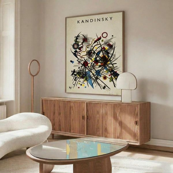 May include: Framed Kandinsky abstract art print hanging above wooden sideboard in modern living room. Artwork features dynamic composition of intersecting black lines, geometric shapes, and vibrant colors including red, blue, yellow on cream background. White upholstered chair and glass coffee table visible in foreground, creating mid-century modern interior styling.