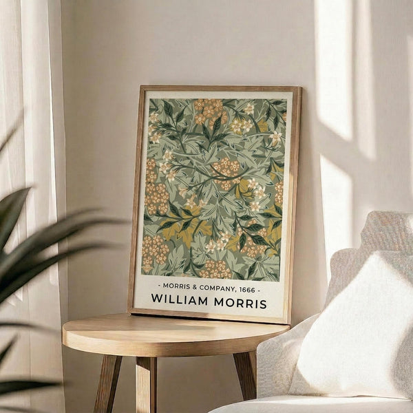 May include: Framed botanical art print with green leafy pattern and small orange flowers displayed on round wooden side table next to beige upholstered chair. Natural sunlight streams across neutral-toned room with plant leaves visible in corner. Print shows decorative floral design in sage green with golden accents in thin wooden frame.