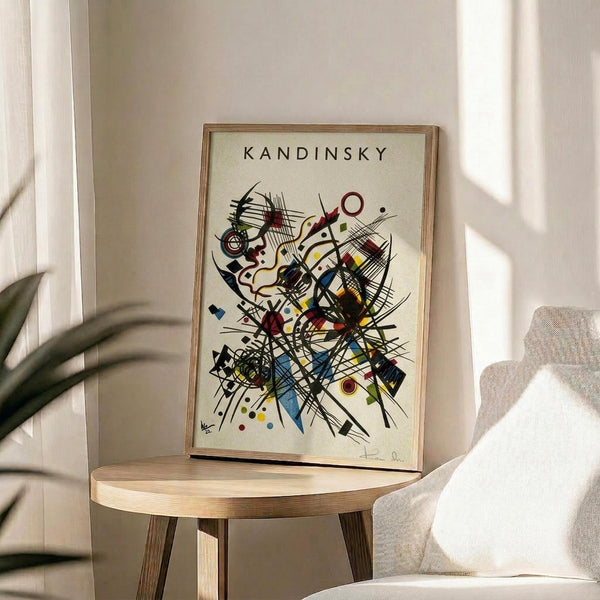May include: Framed abstract art print with geometric shapes, lines, and colorful elements in Kandinsky's distinctive style displayed on a light wood round side table. Cream colored walls with natural sunlight streaming through window, beige upholstered chair partially visible, and green plant leaves in corner create a minimalist modern living space setting.
