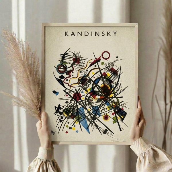 May include: Person holding framed abstract art print with geometric shapes, lines, and colorful forms in Kandinsky's distinctive style. Light wooden frame against cream background with dried pampas grass visible. Features dynamic composition of intersecting black lines, circles, triangles, and organic shapes in blues, reds, yellows, and earth tones on textured cream paper.