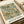 May include: Vintage botanical poster print with intricate green foliage, orange and cream flowers in decorative Arts and Crafts style pattern, displayed in light wood frame on cream colored surface next to neutral fabric roll