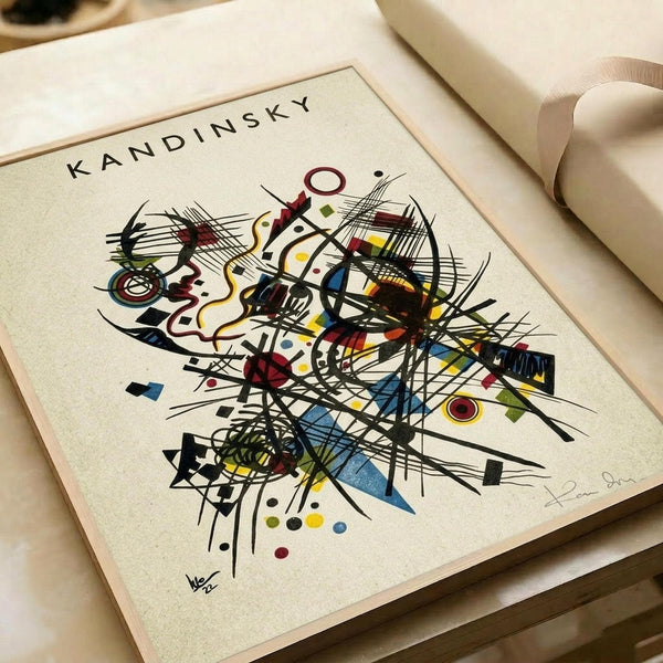 Abstract artwork featuring dynamic black lines intersecting with colorful geometric shapes including circles, triangles, and curved forms in red, blue, yellow, and green. The composition has an energetic, musical quality with flowing lines and scattered dots against a cream background. The print appears on textured paper with "KANDINSKY" text at the top and artist signature at bottom, displayed alongside rolled paper tubes on a light wooden surface.