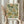 May include: Hands holding a framed botanical art print featuring William Morris-style floral pattern in sage green, orange, and cream colors with ornate leaves and small flowers. Person wears beige ruffle-sleeved top and holds dried pampas grass stems. Soft natural lighting against neutral curtained background creates warm, aesthetic home decor styling scene.