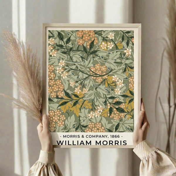 May include: Hands holding a framed botanical art print featuring William Morris-style floral pattern in sage green, orange, and cream colors with ornate leaves and small flowers. Person wears beige ruffle-sleeved top and holds dried pampas grass stems. Soft natural lighting against neutral curtained background creates warm, aesthetic home decor styling scene.