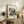 May include: Framed abstract art print with geometric shapes, lines, and circles in vibrant colors including red, blue, yellow, and black on cream background. Light wood frame displayed on rustic wooden sideboard next to potted fern, ceramic vase, and stacked books in neutral-toned living room setting.