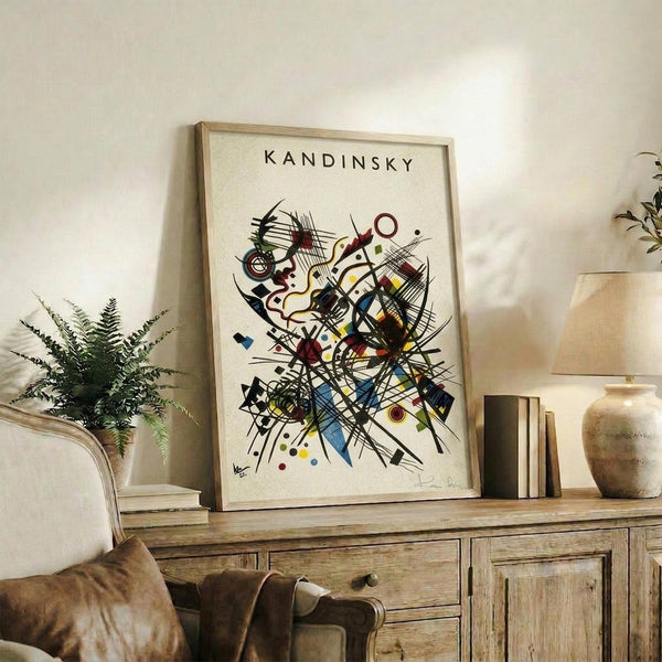 May include: Framed abstract art print with geometric shapes, lines, and circles in vibrant colors including red, blue, yellow, and black on cream background. Light wood frame displayed on rustic wooden sideboard next to potted fern, ceramic vase, and stacked books in neutral-toned living room setting.