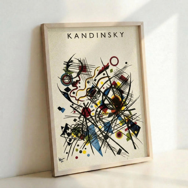 May include: Framed abstract art print in light wood frame leaning against white wall. The artwork features dynamic black lines, geometric shapes, and colorful elements in red, blue, yellow, and green on cream background. "KANDINSKY" text appears at top. Kandinsky's signature visible in bottom left corner. The composition shows energetic movement with intersecting lines, circles, triangles, and organic forms in the artist's distinctive abstract expressionist style.