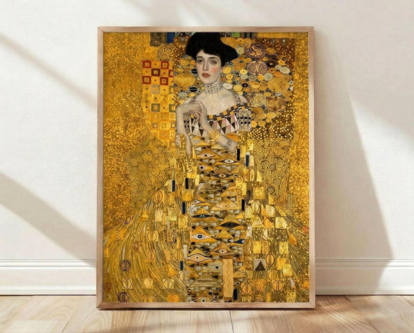 May include: Framed reproduction of Gustav Klimt's golden portrait painting showing an elegant woman in an ornate gold dress with geometric patterns and swirling designs, displayed in a light wooden frame against white walls on wooden flooring with natural sunlight streaming across the scene.