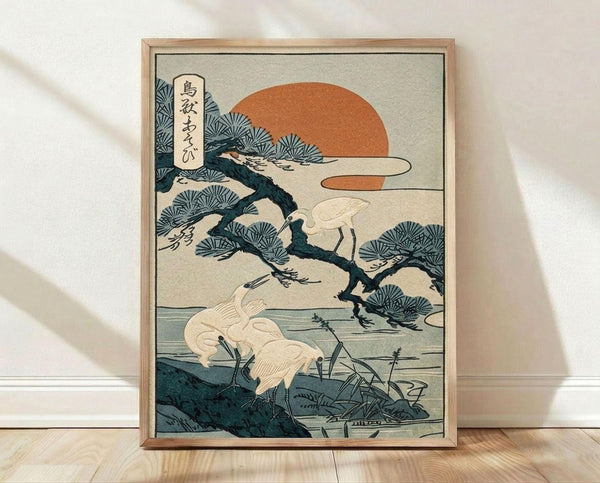 May include: Framed Japanese woodblock print artwork leaning against white wall on light wooden floor. The vintage-style illustration shows white cranes or herons among pine trees beside water, with orange sun in background and vertical Japanese text banner. Artwork has muted teal, orange, and cream colors in traditional ukiyo-e style, displayed in natural wood frame.