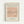 A framed minimalist line drawing of a dachshund dog created with a single continuous black line on a peachy-pink square background. The artwork is displayed in a light wood frame against a cream-colored mat. Text above reads "PICASSO LINE DRAWINGS & PRINTS" and below shows "LE CHIEN" with "Les Métamorphoses, 1930" along with Picasso's signature. The frame hangs on a white wall.