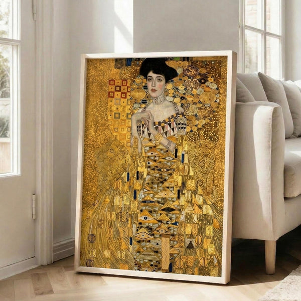 May include: Large framed art print leaning against white wall next to beige sofa in bright living room. The artwork shows a woman with dark hair wearing an ornate golden dress, surrounded by intricate geometric patterns and circular motifs in warm gold, bronze, and yellow tones. White frame contrasts against the predominantly golden artwork. Natural light streams through tall windows, illuminating the modern interior with light wood floors.