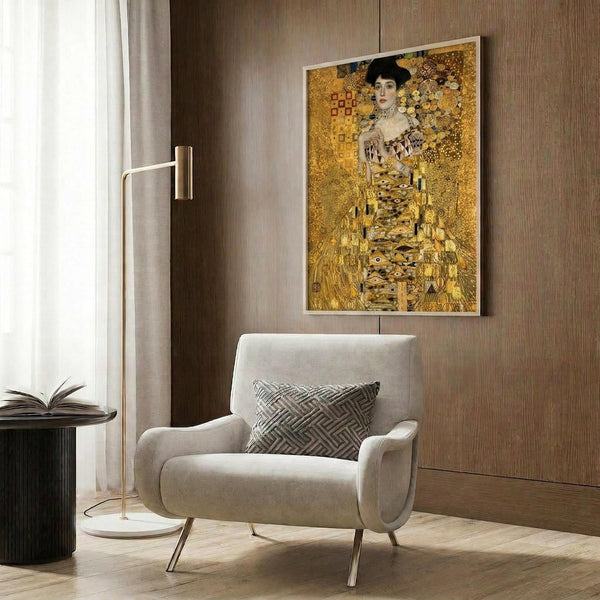 May include: Gustav Klimt's golden portrait of Adele Bloch-Bauer displayed as a framed print on wood-paneled wall above beige armchair with geometric pillow. The artwork features a woman in ornate golden dress with intricate patterns and circles. Modern living room setting with brass floor lamp, dark round side table with open book, and warm neutral tones throughout.