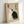 May include: Framed artwork leaning against white wall showing two cats - one white, one black - sitting back-to-back in yin yang formation on textured beige background. Red square seal and black Japanese characters in upper corners. Natural wood frame with shallow depth. Soft lighting creates gentle shadows on wall.
