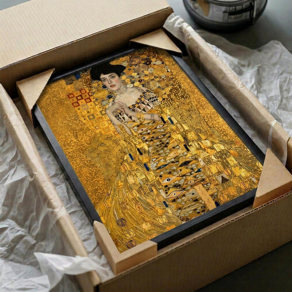 A framed reproduction of Klimt's portrait showing a woman in an ornate golden dress with geometric patterns and swirling designs. The artwork features rich gold, bronze, and amber tones with intricate decorative elements throughout. The black-framed print is carefully packed in a cardboard shipping box with protective tissue paper, sitting on white fabric wrapping materials.