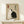 May include: Framed poster with beige background featuring two cats in yin-yang formation - one white cat and one black cat sitting together with tails curved around each other. Japanese characters appear in black ink on the right side with a red seal stamp on the left. The wooden frame sits on a light wood shelf next to a small wrapped white gift box with evergreen sprig. Minimalist room setting with white walls.