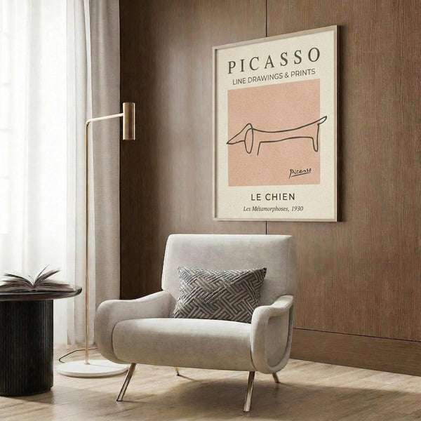 May include: Framed poster with beige background featuring minimalist black line drawing of a dachshund dog, titled "PICASSO LINE DRAWINGS & PRINTS" and "LE CHIEN Les Métamorphoses, 1930". Displayed in modern living room with light gray upholstered chair, geometric patterned pillow, gold floor lamp, dark round side table with open book, and wood panel wall.