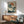May include: Japanese woodblock print artwork featuring white cranes near water with pine trees and orange sun, displayed in wooden frame above mid-century modern walnut credenza. Green upholstered armchair with wooden arms sits nearby, white ceramic table lamp on credenza. Warm sunlight creates geometric shadows on white wall in minimalist living room setting.